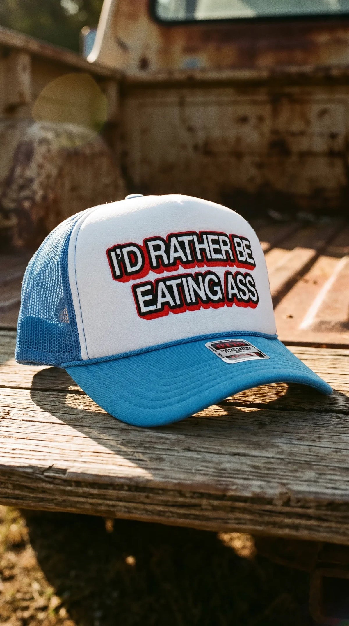 Dirty Merch Apparel blue and white trucker hat with bold I'd Rather Be Eating Ass graphic text on foam front panel