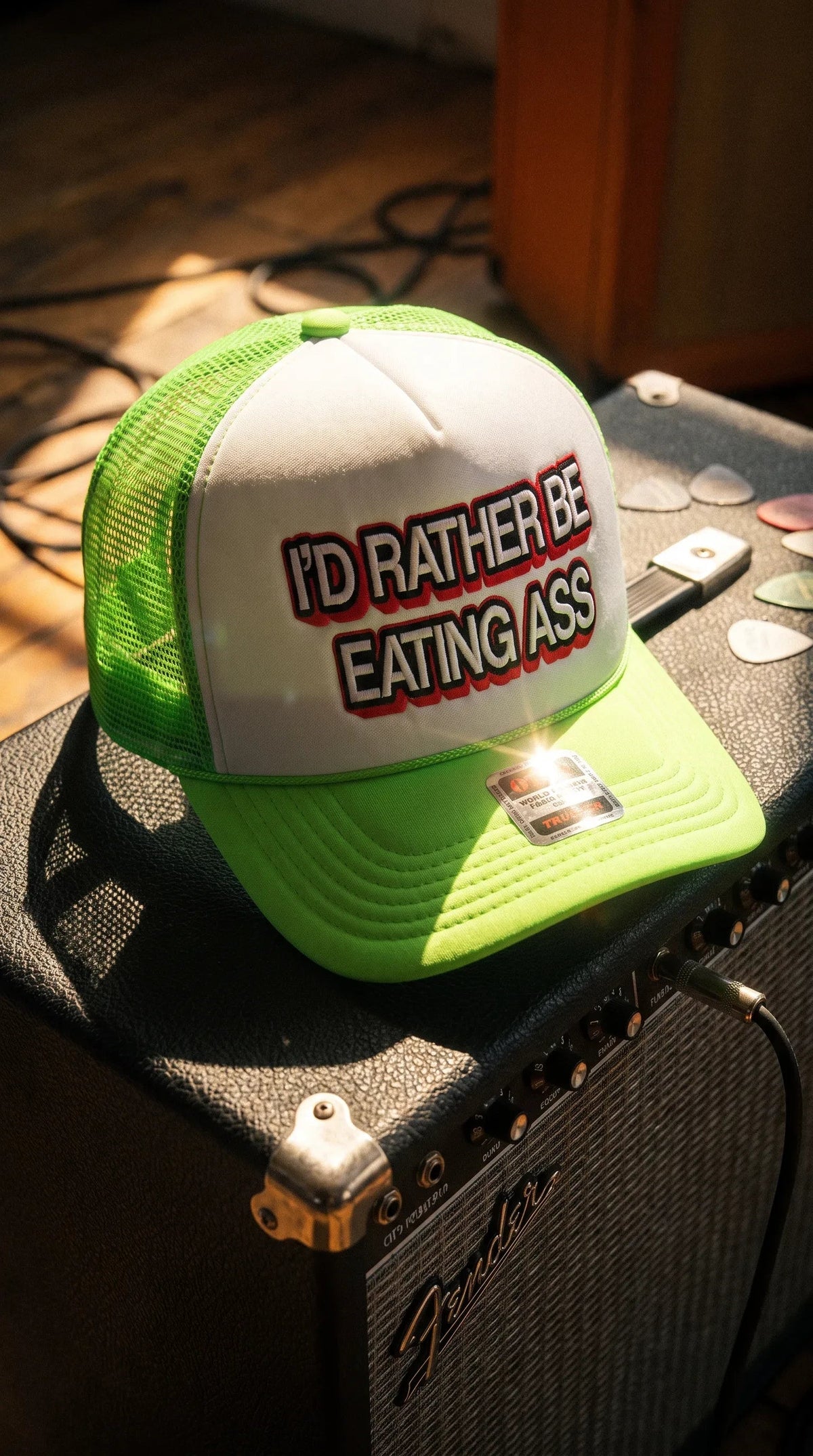 Dirty Merch Apparel neon green and white trucker hat with bold text graphic sitting on a guitar amplifier